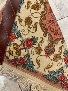 Azerbaijan Tappeti Milano Un tappeto dettagliato e colorato con motivi floreali, bordo sfrangiato e un angolo in pelle su un pavimento piastrellato.