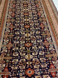 Azerbaijan Tappeti Milano Intricato tappeto persiano con motivi geometrici e floreali, colori prevalentemente scuri e un logo a foglie multicolore.