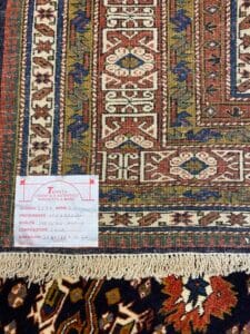 Azerbaijan Tappeti Milano Primo piano di un tappeto orientale a fantasia con un'etichetta in italiano e un logo a forma di foglia colorata nell'angolo.