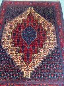 Azerbaijan Tappeti Milano Tappeto in stile persiano con medaglione centrale a rombi, motivi rossi e blu e un logo a forma di foglia colorata in un angolo.