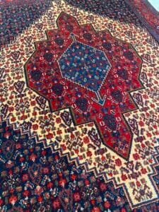 Azerbaijan Tappeti Milano Tappeto persiano con motivi geometrici e floreali rossi, blu e crema e un logo a forma di foglia colorata.