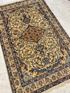 Azerbaijan Tappeti Milano Un tappeto persiano a motivi beige e blu con intricati disegni floreali giace su un pavimento piastrellato chiaro.