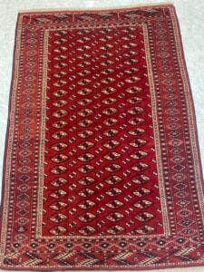 Azerbaijan Tappeti Milano Tappeto a fantasia rossa e marrone con disegni geometrici, delimitati da motivi intricati, su un pavimento piastrellato chiaro.