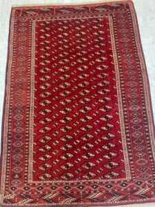 Azerbaijan Tappeti Milano Un tappeto rosso con disegni geometrici e un campione di filato colorato attaccato nell'angolo in alto a destra.