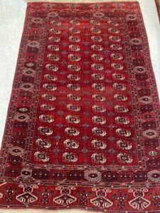 Azerbaijan Tappeti Milano Un tappeto rosso con disegni geometrici, esposto su un pavimento piastrellato, e un logo a forma di foglia colorata in un angolo.