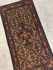 Azerbaijan Tappeti Milano Tappeto persiano a motivi colorati su un pavimento piastrellato, con un logo floreale multicolore nell'angolo in alto a destra.