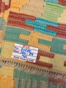Azerbaijan Tappeti Milano Primo piano di un tappeto colorato con motivi geometrici e un'etichetta che recita: "IPAK, No: 50584, Dimensioni: 180 x 74, 161, Fatto a mano in Iran". Nell'angolo in alto a destra è visibile il logo di una foglia multicolore.