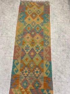 Azerbaijan Tappeti Milano Tappeto lungo e rettangolare con motivi geometrici nei toni dell'arancio, del blu, del verde e del beige, posto su un pavimento piastrellato grigio chiaro. Un logo artistico colorato si trova nell'angolo in alto a destra.