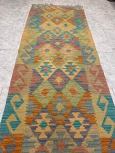 Azerbaijan Tappeti Milano Un lungo tappeto rettangolare con motivi geometrici nei toni della terra, verde, arancione, giallo e blu, esposto su un pavimento piastrellato. Un logo colorato con una "A" si trova nell'angolo in alto a destra.