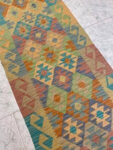 Azerbaijan Tappeti Milano Un tappeto colorato, a motivi geometrici, con forme a rombi e chevron nei toni della terra, verde, blu, arancione, giallo e beige, posato su un pavimento di piastrelle chiare. Un logo floreale multicolore si trova in alto a destra.