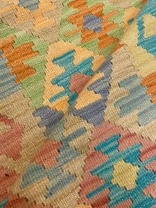 Azerbaijan Tappeti Milano Primo piano di un tessuto colorato a motivi geometrici con forme a zig zag nei toni del blu, verde, arancione e rosa. Nell'angolo in alto a destra è presente un piccolo logo a forma di foglia multicolore.