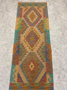 Azerbaijan Tappeti Milano Tappeto rettangolare con motivi geometrici a rombi e a zig-zag nei toni della terra, marrone, arancione, verde, giallo e blu, esposto su un pavimento piastrellato grigio chiaro. Nell'angolo in alto a destra compare il logo "A" colorato.