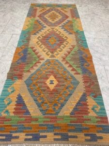 Azerbaijan Tappeti Milano Un lungo tappeto rettangolare intrecciato con motivi geometrici nei toni della terra, marrone, arancione, blu, verde e giallo, esposto su un pavimento piastrellato chiaro. Un logo colorato si trova nell'angolo in alto a destra.