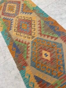 Azerbaijan Tappeti Milano Un tappeto colorato con disegni geometrici a rombi nei toni della terra, verde, blu e arancione, giace su un pavimento piastrellato grigio chiaro. Nell'angolo in alto a destra compare una "A" decorativa con il logo di una foglia colorata.