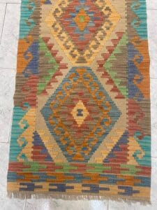 Azerbaijan Tappeti Milano Tappeto rettangolare con motivi geometrici a rombi e a zig zag nei toni della terra, blu, giallo, arancione, verde e rosso, esposto su un pavimento di piastrelle chiare. Un logo colorato si trova nell'angolo in alto a destra.