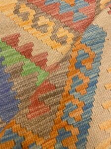 Azerbaijan Tappeti Milano Primo piano di un tessuto colorato a motivi geometrici con forme a zig-zag e a rombi nei toni del blu, verde, rosso, giallo e beige, con una foglia multicolore e il logo "A" stilizzato nell'angolo.