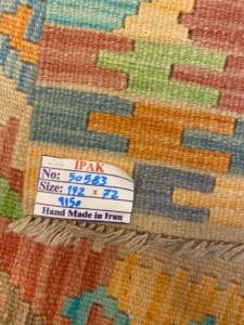 Azerbaijan Tappeti Milano Primo piano di un tappeto colorato con etichetta "IPAK, No: 50583, Size: 192 x 72, 9156, Hand Made in Iran". Il tappeto presenta motivi geometrici in tonalità pastello.