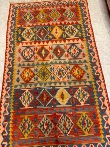 Azerbaijan Tappeti Milano Tappeto kilim rettangolare con motivi geometrici a rombi nei toni del rosso, giallo, verde, blu e beige, delimitati da motivi a zig zag. Il tappeto è posato su un pavimento piastrellato chiaro.