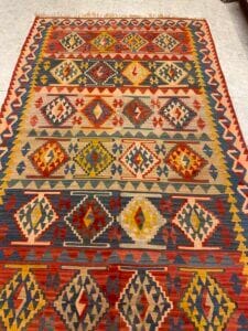 Azerbaijan Tappeti Milano Tappeto colorato con disegni geometrici a rombi e a zig-zag nei toni del rosso, del giallo, del blu e del beige, steso su un pavimento piastrellato chiaro. Nell'angolo superiore destro dell'immagine compare una farfalla.