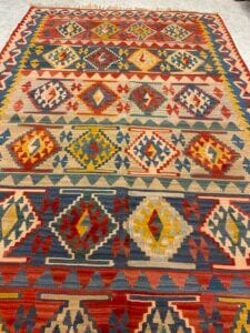 Azerbaijan Tappeti Milano Tappeto rettangolare colorato con motivi geometrici in rosso, blu, giallo e beige, caratterizzato da file di rombi e triangoli. Nell'angolo in alto a destra compare il logo di una piccola farfalla multicolore.