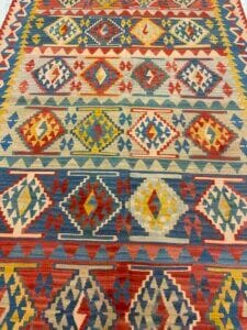 Azerbaijan Tappeti Milano Tappeto intrecciato con motivi geometrici in rosso, blu, giallo e beige. Il tappeto presenta motivi a rombi e triangoli ripetuti, con un logo a forma di foglia colorata nell'angolo in alto a destra.