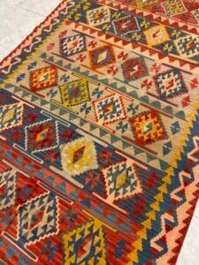 Azerbaijan Tappeti Milano Un tappeto colorato a motivi geometrici con disegni a rombi e a zig zag nei toni del rosso, del giallo, del blu e del verde giace su un pavimento piastrellato. In un angolo è visibile un logo multicolore con una firma.