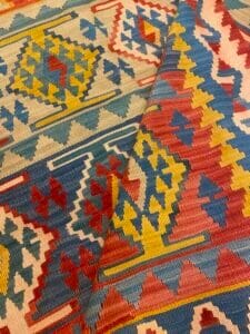 Azerbaijan Tappeti Milano Primo piano di un tappeto colorato con intricati motivi geometrici in rosso, blu, giallo e crema. Nell'angolo in alto a destra compare la grafica di una farfalla con ali rosse, gialle, blu e verdi.