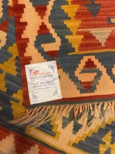 Azerbaijan Tappeti Milano Primo piano di un tappeto azero colorato con motivi geometrici in rosso, blu e giallo. Un cartellino con dettagli scritti a mano è attaccato alla frangia e un logo a forma di foglia colorata appare nell'angolo in alto a destra.