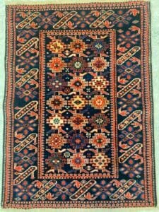 Azerbaijan Tappeti Milano Tappeto rettangolare con intricati motivi geometrici nei toni del blu, dell'arancio, del crema e del rosso, caratterizzato da un pannello centrale con motivi a stella e da un bordo decorativo. Il tappeto è posato su un pavimento piastrellato.