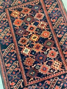 Azerbaijan Tappeti Milano Un tappeto colorato e fantasia presenta motivi geometrici nei toni del rosso, del blu e del crema, con un bordo audace di foglie e forme floreali. Una rosa dei venti multicolore è posizionata nell'angolo in alto a destra.