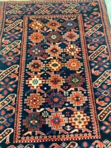 Azerbaijan Tappeti Milano Tappeto dalla ricca fantasia con disegni geometrici in rosso, arancione, crema e verde su uno sfondo blu scuro. Un logo colorato a forma di foglia con un cuore è visibile nell'angolo in alto a destra.