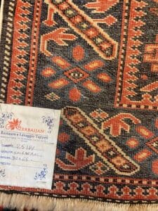 Azerbaijan Tappeti Milano Primo piano di un tappeto a motivi geometrici rossi, neri e beige. Nell'angolo in basso a sinistra è applicata un'etichetta con testo e logo a foglie colorate di "Azerbaijan Restauro e Lavaggio Tappeti".