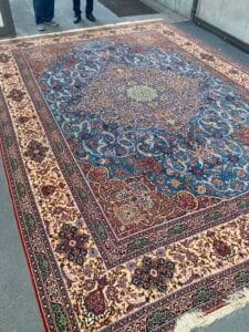 Azerbaijan Tappeti Milano Tappeto persiano decorato blu e rosso con intricati motivi floreali in mostra. Due persone e una ruota di bicicletta sono parzialmente visibili sullo sfondo.