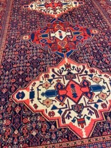 Azerbaijan Tappeti Milano Tappeto tradizionale in stile persiano con intricati motivi geometrici in rosso, blu e crema, con medaglioni centrali e motivi floreali. Nell'angolo in alto a destra è visibile il logo di una foglia colorata.