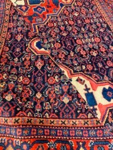 Azerbaijan Tappeti Milano Primo piano di un tappeto persiano a motivi colorati con intricati disegni rossi, blu e arancioni. Il tappeto è leggermente piegato in diagonale e rivela i dettagliati motivi floreali e geometrici. Nell'angolo compare il logo di una piccola foglia multicolore.