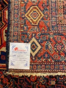 Azerbaijan Tappeti Milano Primo piano di un tappeto a motivi geometrici in rosso, blu e beige. Un'etichetta in italiano con bordi floreali è attaccata, con la scritta "Azerbaijan" e i dettagli sul restauro e la pulizia.