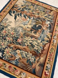 Azerbaijan Tappeti Milano Tappeto decorativo con scena naturalistica con due cigni in uno stagno, circondati da verde lussureggiante, alberi e fiori colorati, delimitati da un motivo floreale. Il tappeto è steso su un pavimento piastrellato.