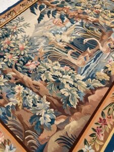 Azerbaijan Tappeti Milano Un arazzo colorato e intricato raffigura una scena lussureggiante e frondosa con cigni che nuotano sull'acqua, circondati da fiori, alberi e fogliame, delimitati da una cornice decorativa.