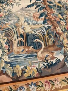 Azerbaijan Tappeti Milano Un arazzo dettagliato raffigura due cigni che nuotano in uno stagno circondato da un fogliame rigoglioso, fiori e alberi, con un paesaggio e un edificio sullo sfondo. Nell'angolo in alto a destra è presente il logo di una farfalla colorata.