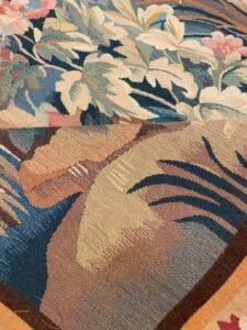 Azerbaijan Tappeti Milano Primo piano di un arazzo colorato con motivi floreali e forme astratte nei toni del blu, beige, rosa e verde. Nell'angolo in alto a destra compaiono una foglia multicolore e la lettera "A".