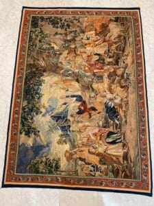 Azerbaijan Tappeti Milano Un dettagliato arazzo tessuto che raffigura una vivace scena all'aperto con numerosi personaggi in abiti storici, circondati da alberi e montagne, bordati da un ornato motivo floreale.