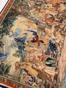 Azerbaijan Tappeti Milano Un arazzo dettagliato e colorato raffigura una scena storica o biblica con molte figure in abiti fluenti, ambientate all'aperto sotto alberi con motivi ornamentali che bordano il tessuto. Nell'angolo in alto a destra compare un moderno logo multicolore.