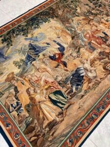 Azerbaijan Tappeti Milano Un arazzo dettagliato e colorato che raffigura una scena storica o biblica con più figure in abito, che interagiscono sotto un albero, bordato da motivi ornamentali. Nell'angolo è visibile un logo con una foglia e una "A".