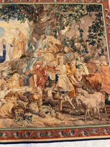 Azerbaijan Tappeti Milano Un arazzo riccamente dettagliato raffigura una scena pastorale con abitanti del villaggio che vanno a prendere l'acqua, accudiscono gli animali e interagiscono vicino a un ruscello sotto gli alberi, con bordi colorati e motivi intricati.