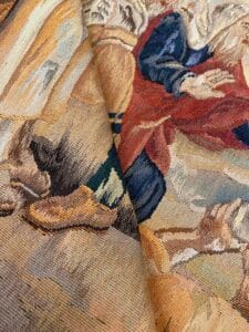 Azerbaijan Tappeti Milano Primo piano di un arazzo piegato e colorato che mostra dettagliati disegni di persone in abiti storici, con mani e scarpe visibili e un logo luminoso nell'angolo in alto a destra.