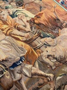 Azerbaijan Tappeti Milano Primo piano di un arazzo dettagliato che raffigura persone e bestiame, tra cui mucche e pecore, nei toni della terra. Nell'angolo in alto a destra compare un logo astratto e colorato con la lettera "A".