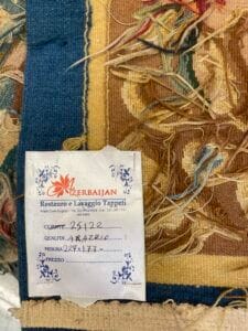 Azerbaijan Tappeti Milano Primo piano di un tappeto parzialmente restaurato con fili sfilacciati, che mostra un'etichetta in italiano che include il nome "Azerbaijan", la qualità, le dimensioni e un prezzo parzialmente compilato, con un logo a foglia colorata nell'angolo in alto a destra.