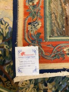 Azerbaijan Tappeti Milano Primo piano di un tappeto colorato con motivi floreali e di foglie. Un cartellino di carta etichettato "Azerbaijan" include dettagli scritti a mano sul restauro e la pulizia del tappeto, con dimensioni annotate di 232x216.