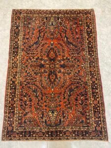 Azerbaijan Tappeti Milano Tappeto persiano rettangolare e decorato con un intricato motivo floreale e geometrico nei toni del rosso, arancio, blu e crema. Il tappeto ha un bordo decorativo ed è esposto su un pavimento piastrellato chiaro.