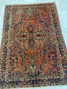 Azerbaijan Tappeti Milano Tappeto persiano rettangolare a motivi intricati nei colori rosso, blu e beige, con disegni floreali e geometrici simmetrici, delimitato da un bordo decorativo scuro, esposto su un pavimento piastrellato chiaro.